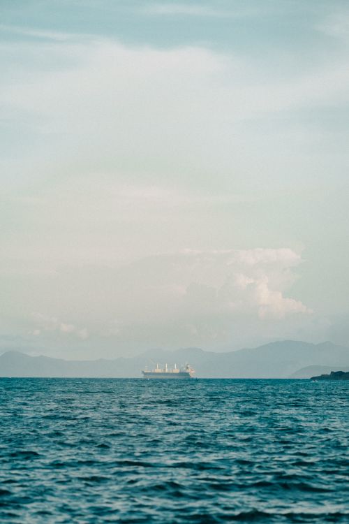 Free Peaceful sea view with a ship on the horizon against mountains under a soft blue sky. Stock Photo