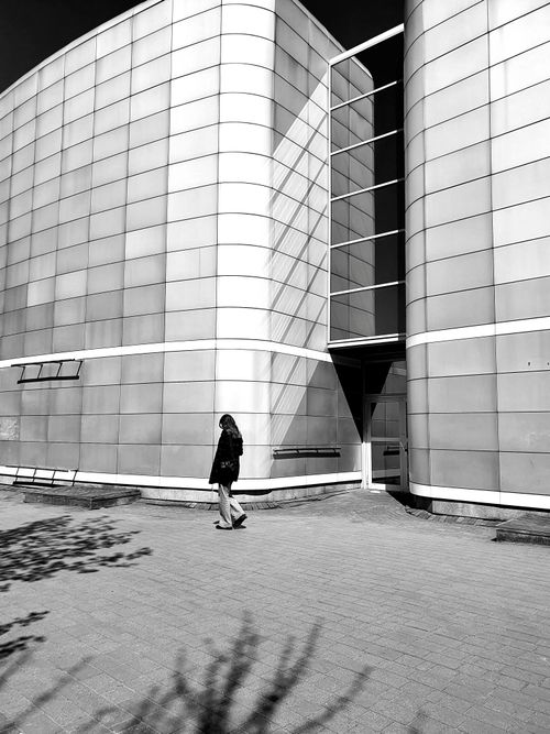 Free Black and white photo of a woman walking past a modern, futuristic building facade. Stock Photo