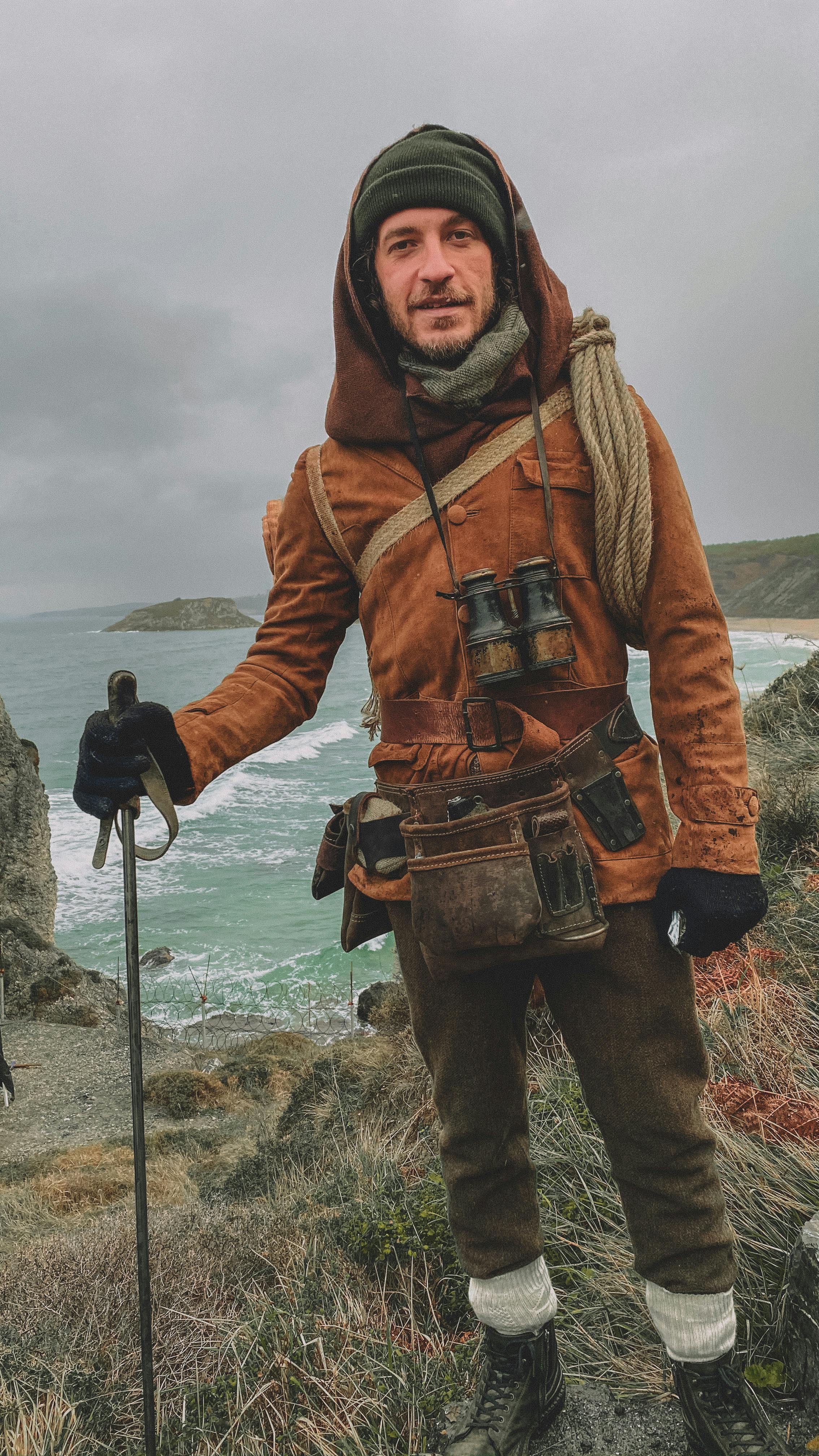 Explorer stands against a rugged coastal backdrop, equipped for a cold adventure with gear and binoculars.