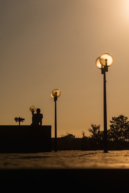 Street Lamps and Silhouette of Man Sitting on Wall at Sunset