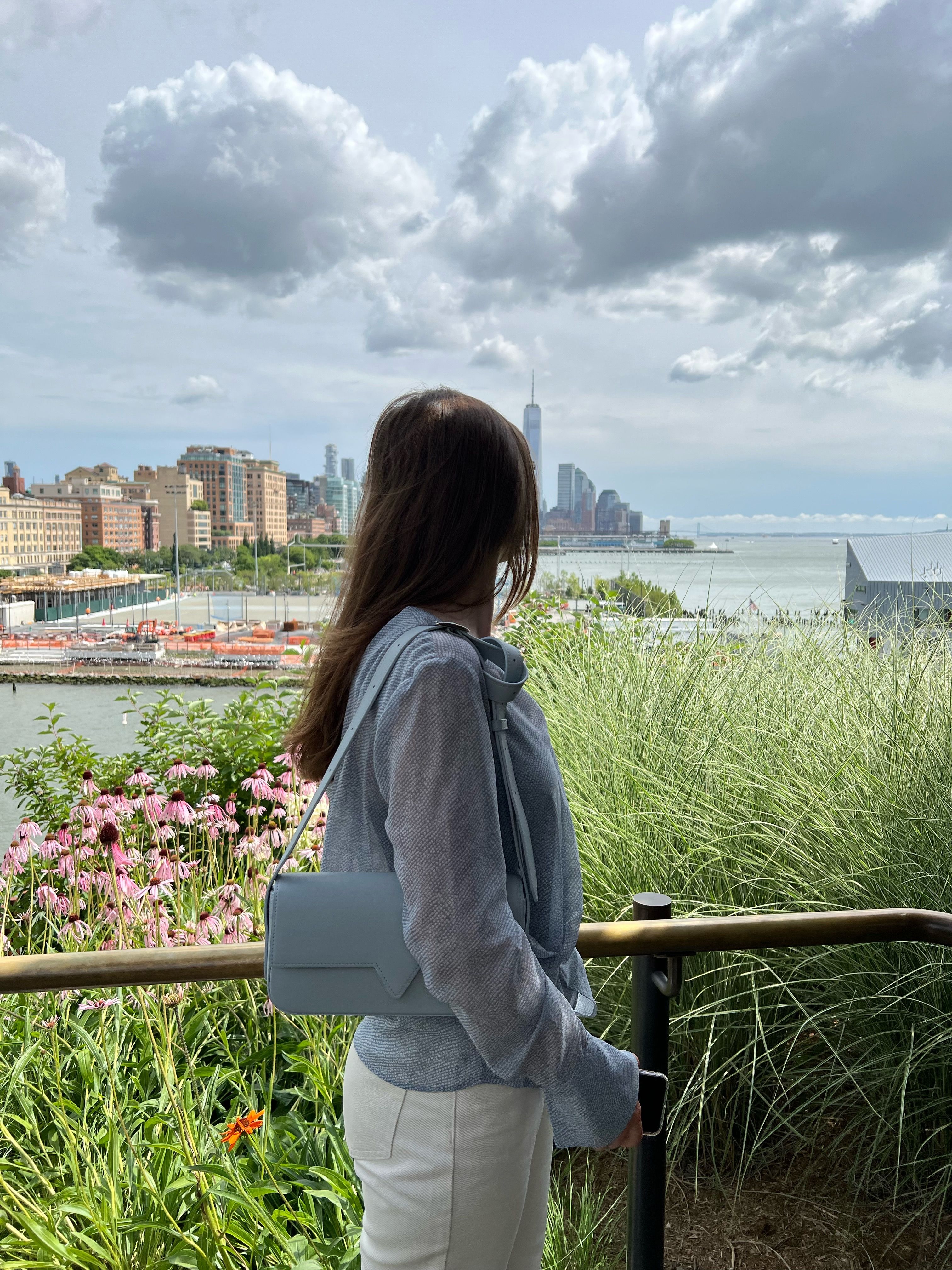 A woman stands by railing, overlooking the iconic New York City skyline and the Hudson River.