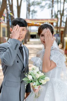 Bride and groom playfully pose outdoors, covering one eye with a hand.