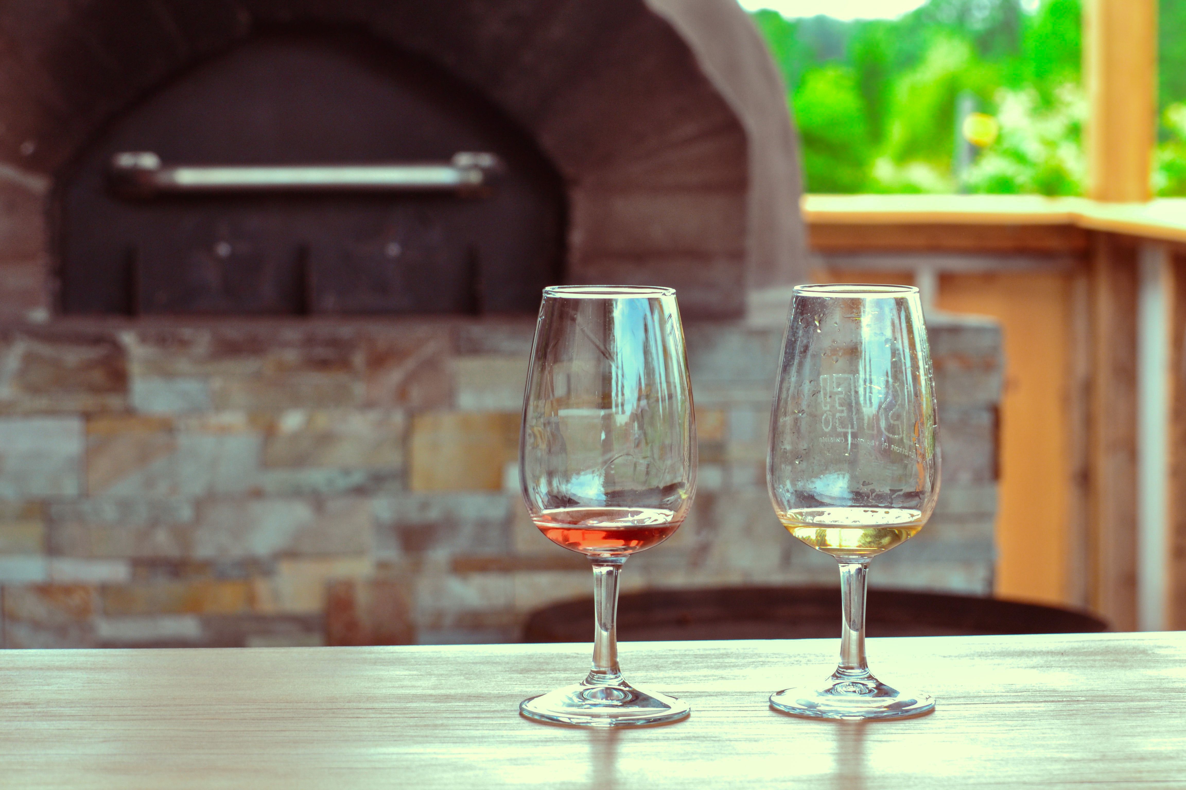 Two empty wine glasses on a table in an outdoor setting with a stone oven in the background.
