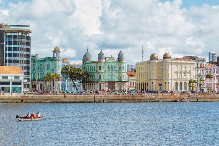 The colorful colonial waterfront of Recife Antigo, with pastel-hued historic buildings reflected on the water and a small boat in the foreground.
