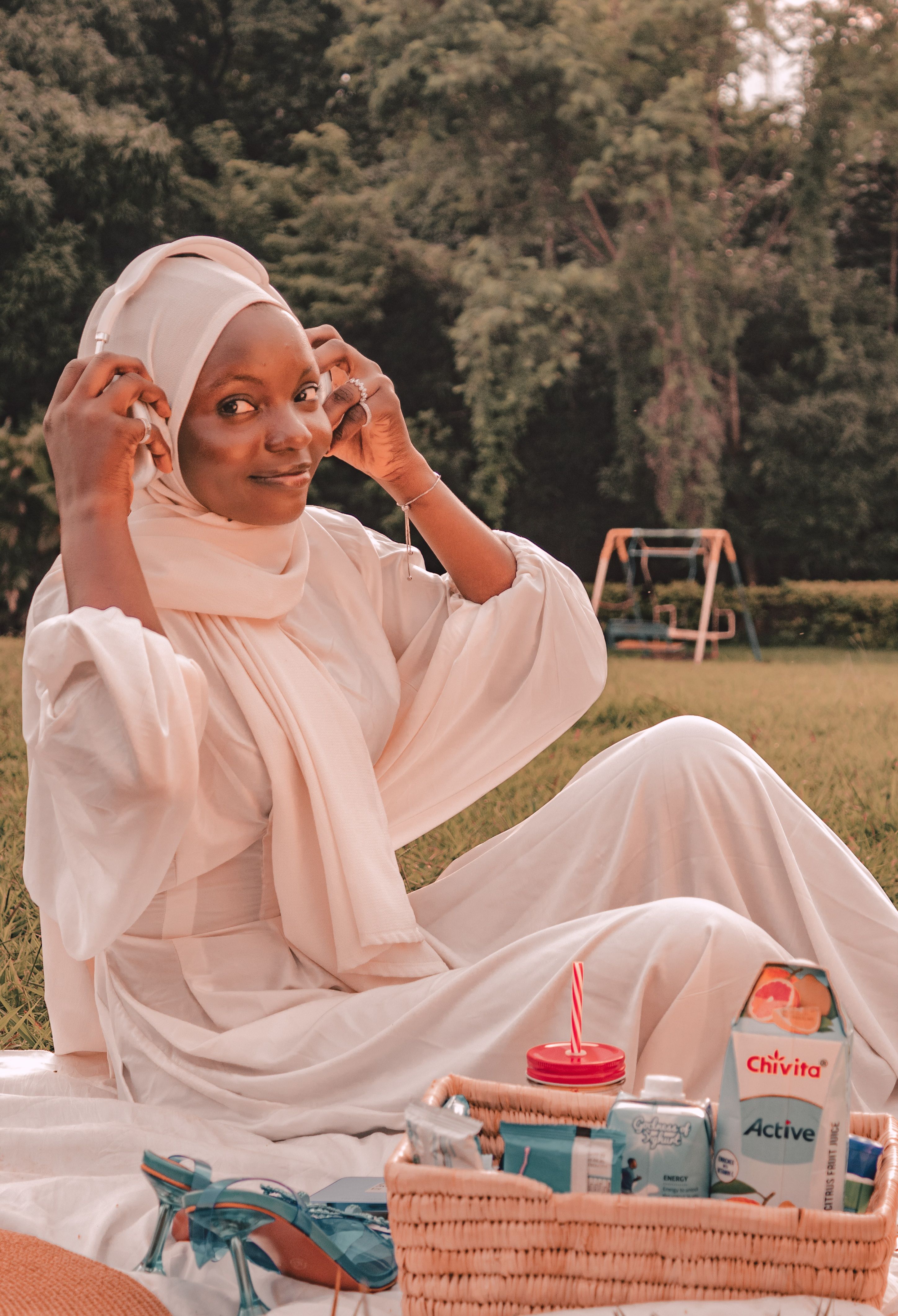 A woman in a hijab relaxes at a picnic in a Nigerian park, enjoying music and refreshments.