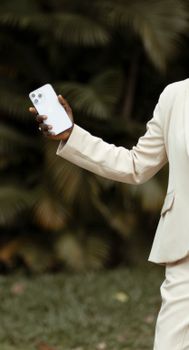 Woman in a white suit holding a smartphone in a park setting.