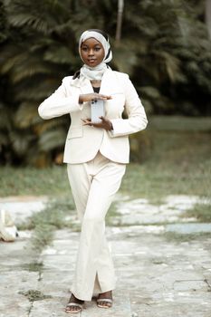 A fashionable woman in a white suit poses elegantly with a smartphone outdoors.