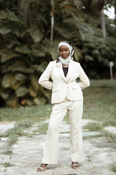 Elegant woman in white suit and headscarf poses in stylish business attire outdoors.