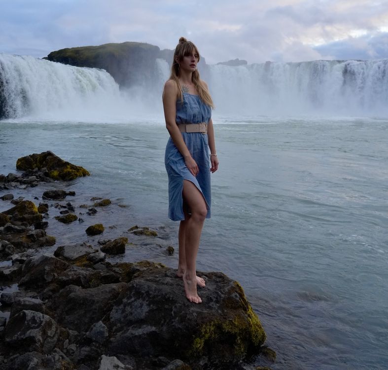 A woman in a blue dress standing on rocks near a waterfall · Free Stock ...