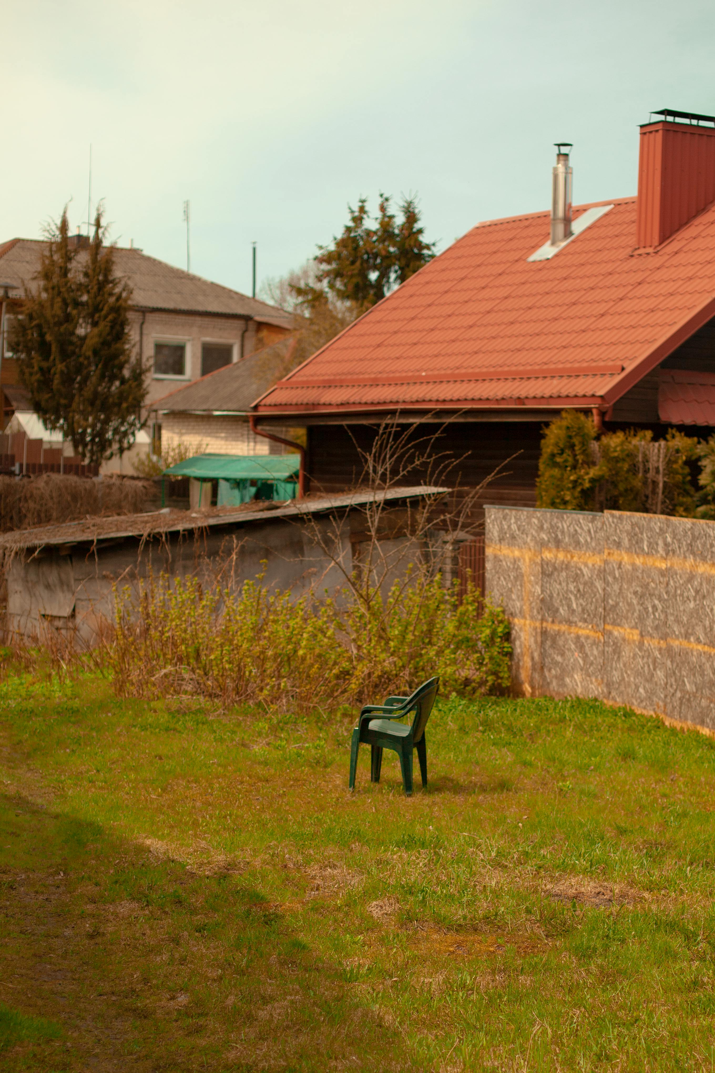 A serene backyard scene featuring a single plastic chair on grass beside village houses.