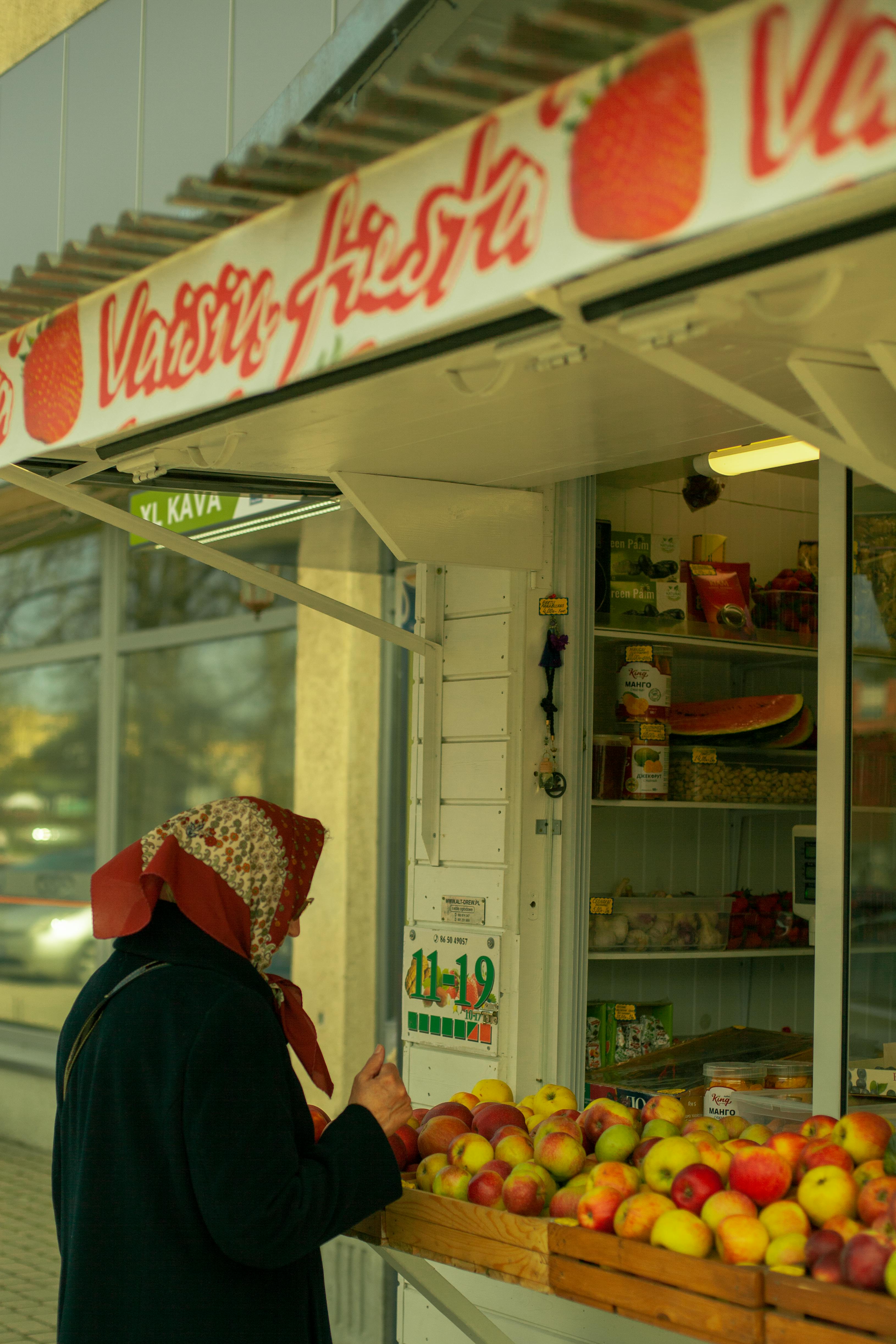 Elderly woman with headscarf buys fresh apples at an urban market stall.