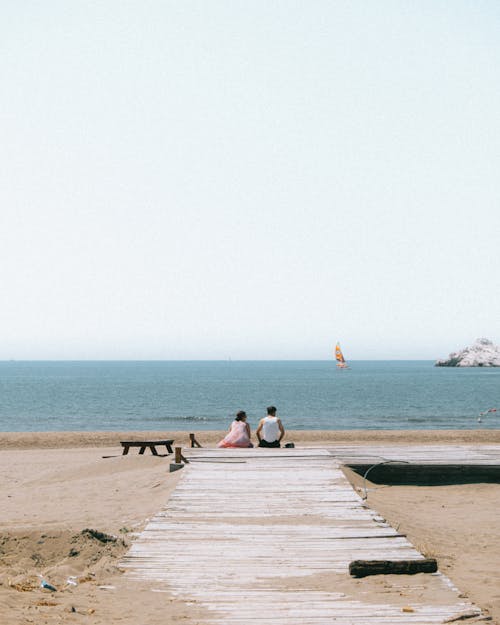 Two People Sitting at the End of a Boardwalk on a Beach 