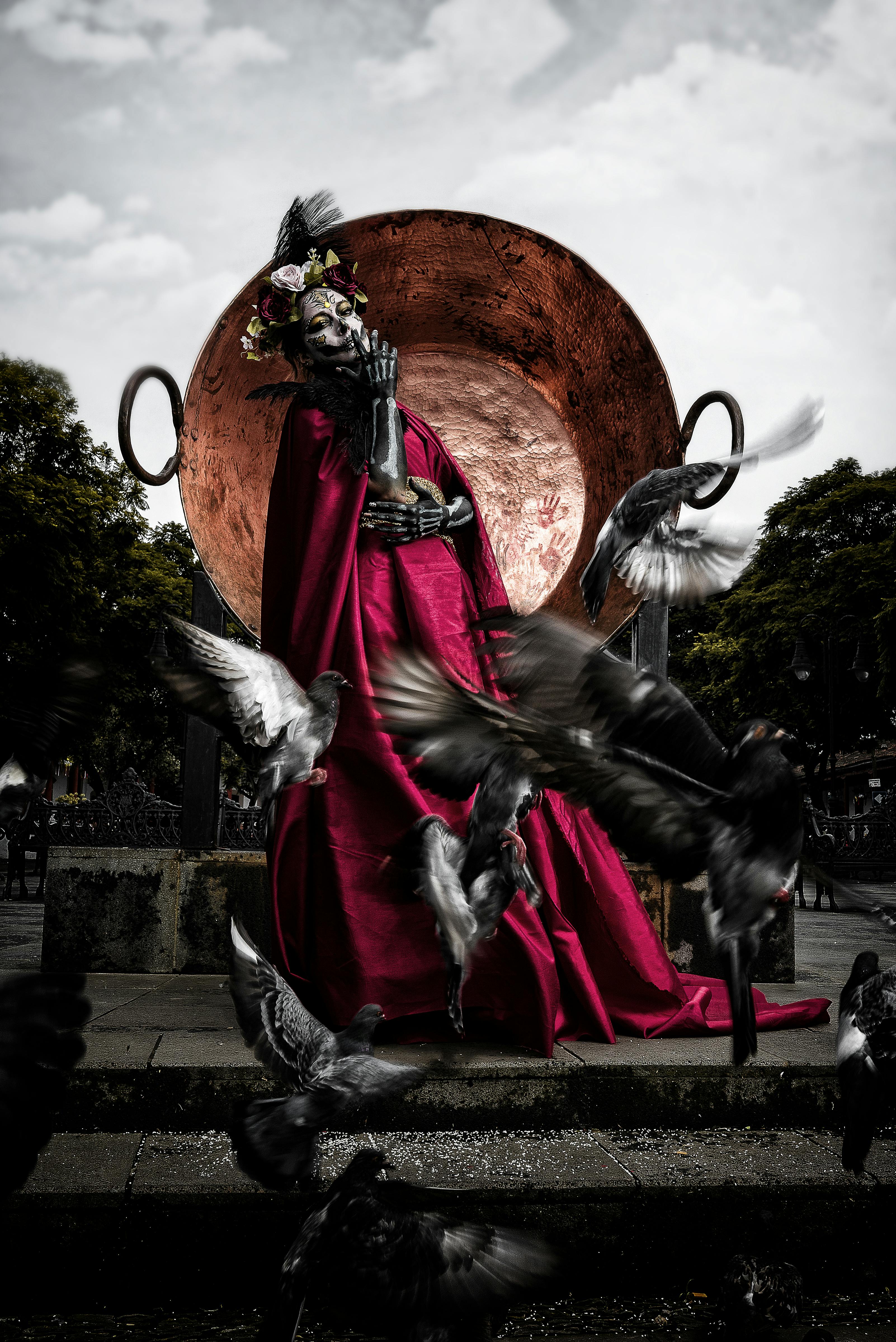 flock of pigeons flying in front of a model in a catrina costume in front of a monument of a huge brass bowl