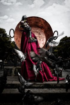 A figure dressed as La Calavera Catrina in a red costume surrounded by pigeons in a park.