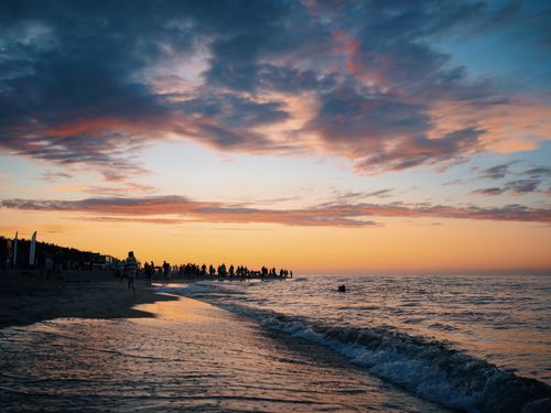 Free A picturesque sunset at the beach with a crowd silhouetted against the vivid sky, waves gently lapping the shore. Stock Photo