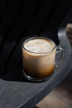 Close-up of iced coffee with foam in a glass mug on a textured dark wood surface.