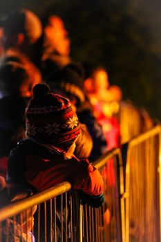 A young boy in a warm hat watches a festival at night, leaning on a barricade.