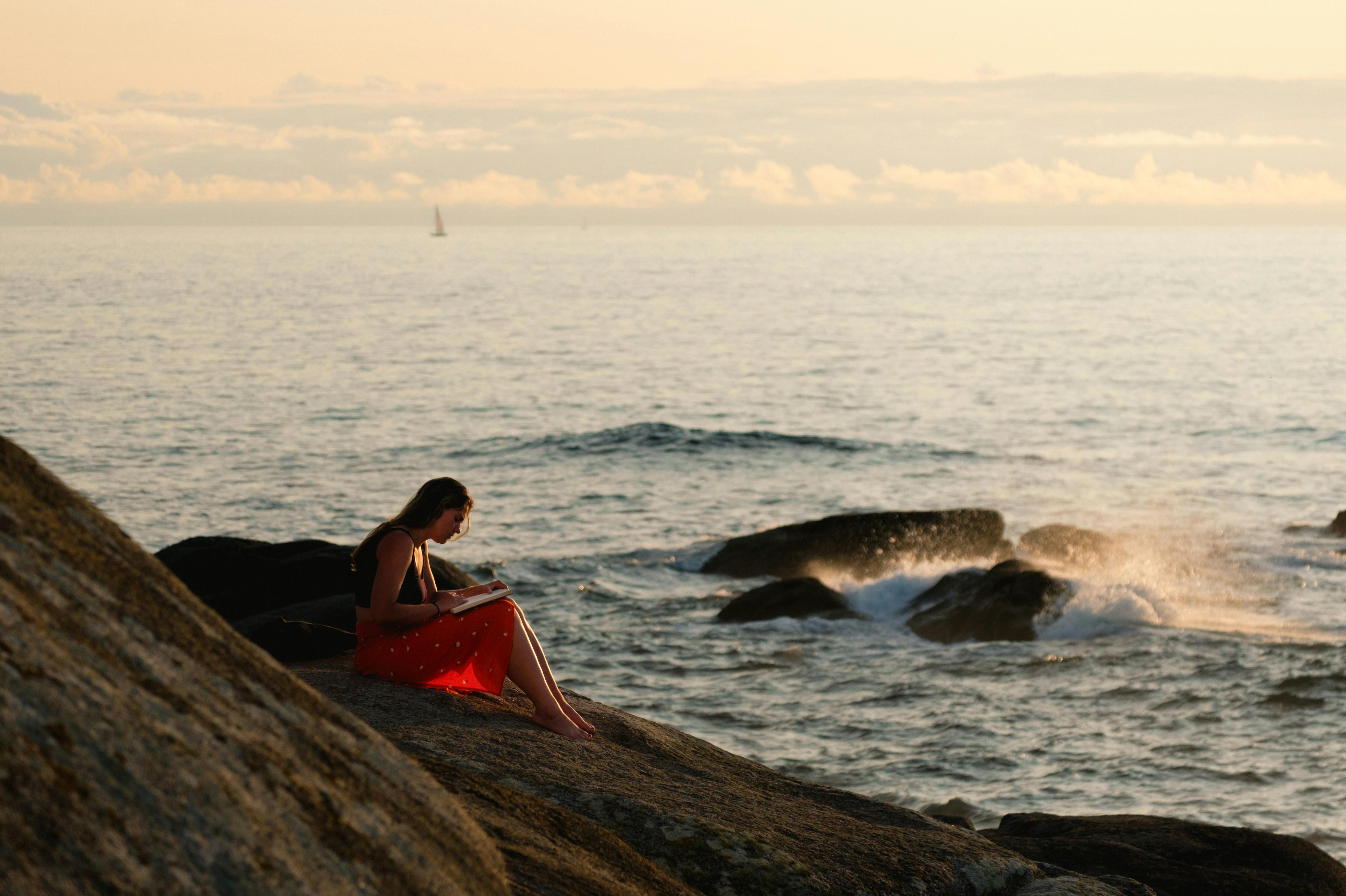 A woman enjoys reading a book by the ocean during a serene sunset.