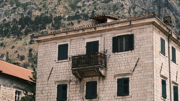 A historic stone building with green window shutters and a balcony, nestled in a mountainous area.