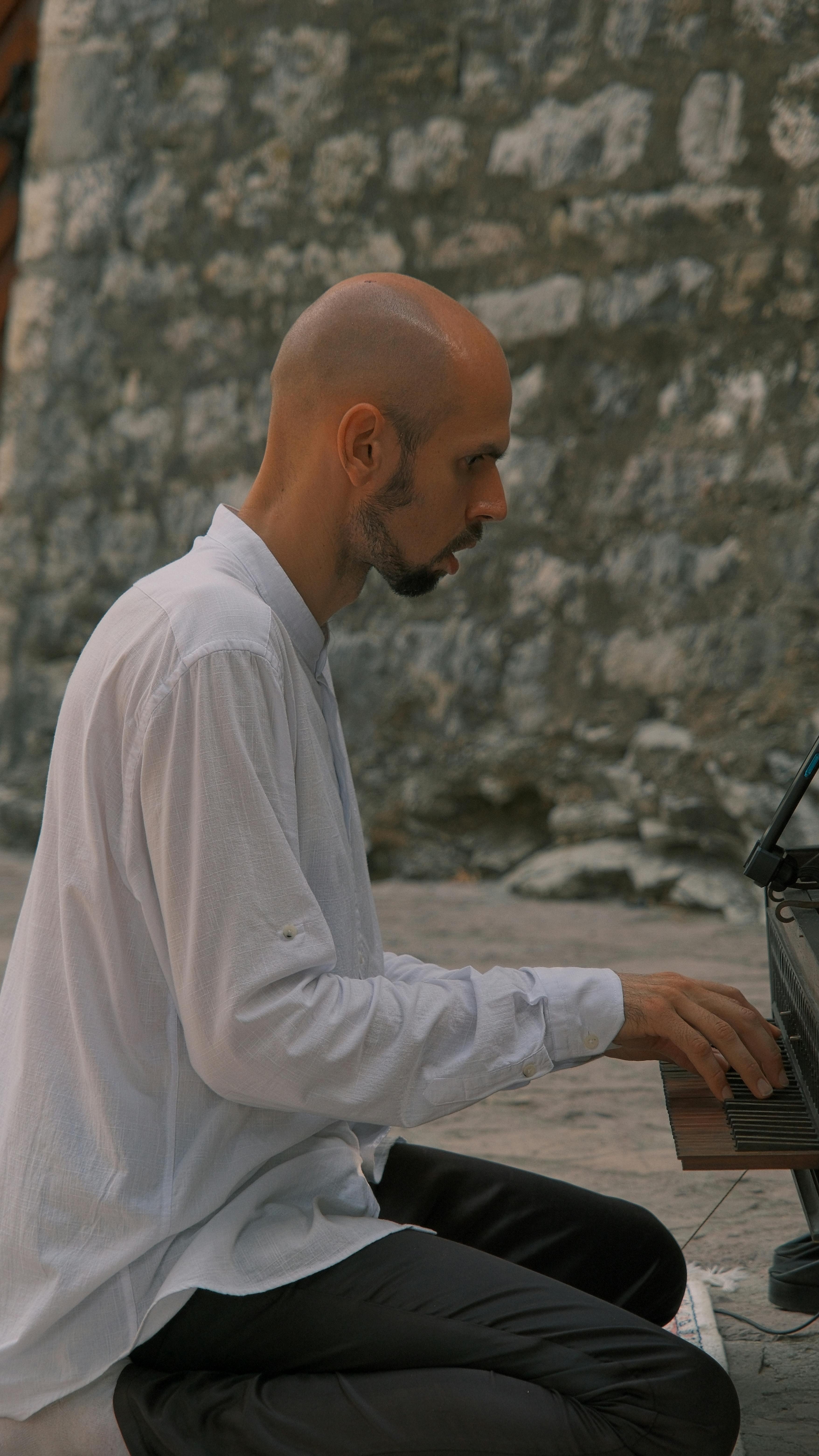 Bald man playing piano outdoors against a stone wall backdrop.