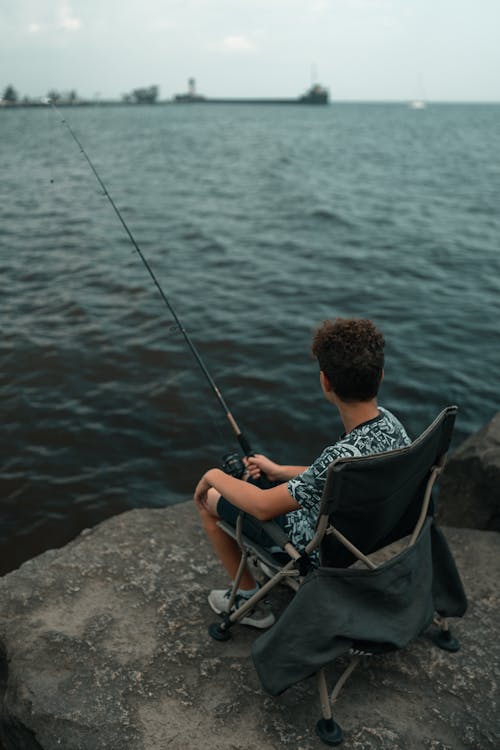 A boy sitting in a chair on the edge of the water with a fishing rod