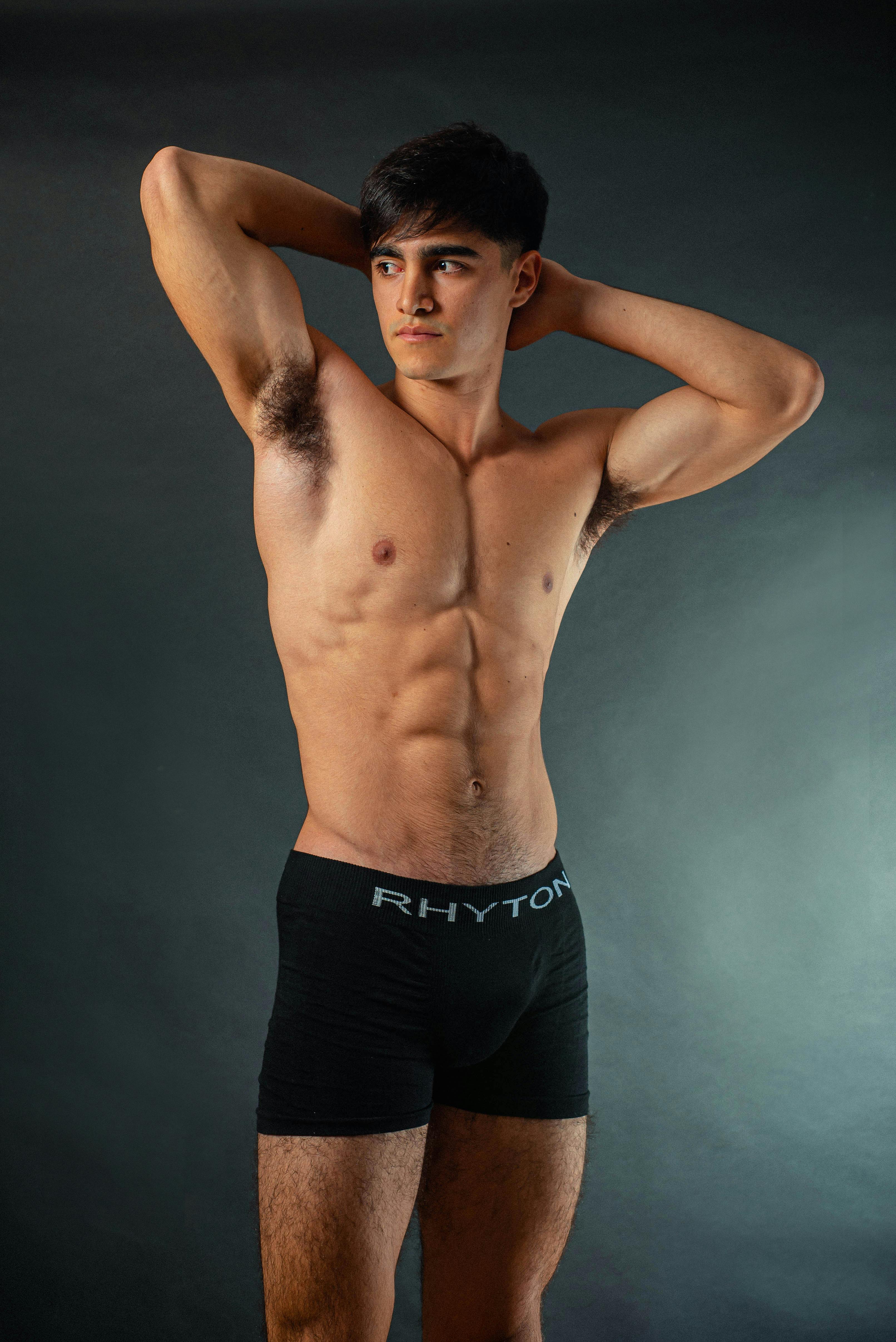Muscular young man posing confidently in underwear against a gray backdrop, showcasing fitness and strength.