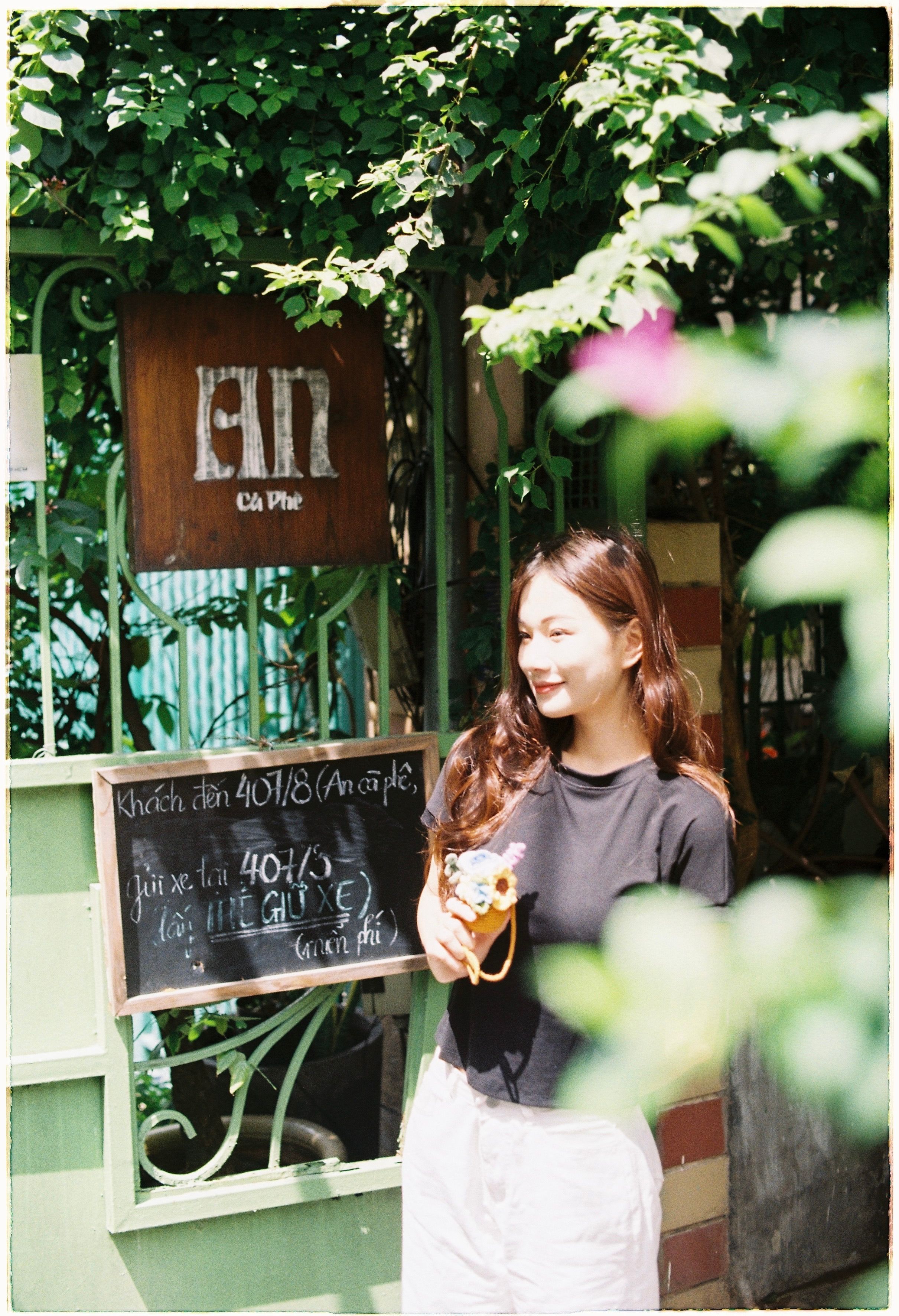 Asian woman with long hair standing beside a blackboard at a sunlit cafe entrance.