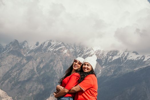 Two women in red shirts and white beanies embracing and smiling against Auli mountains.