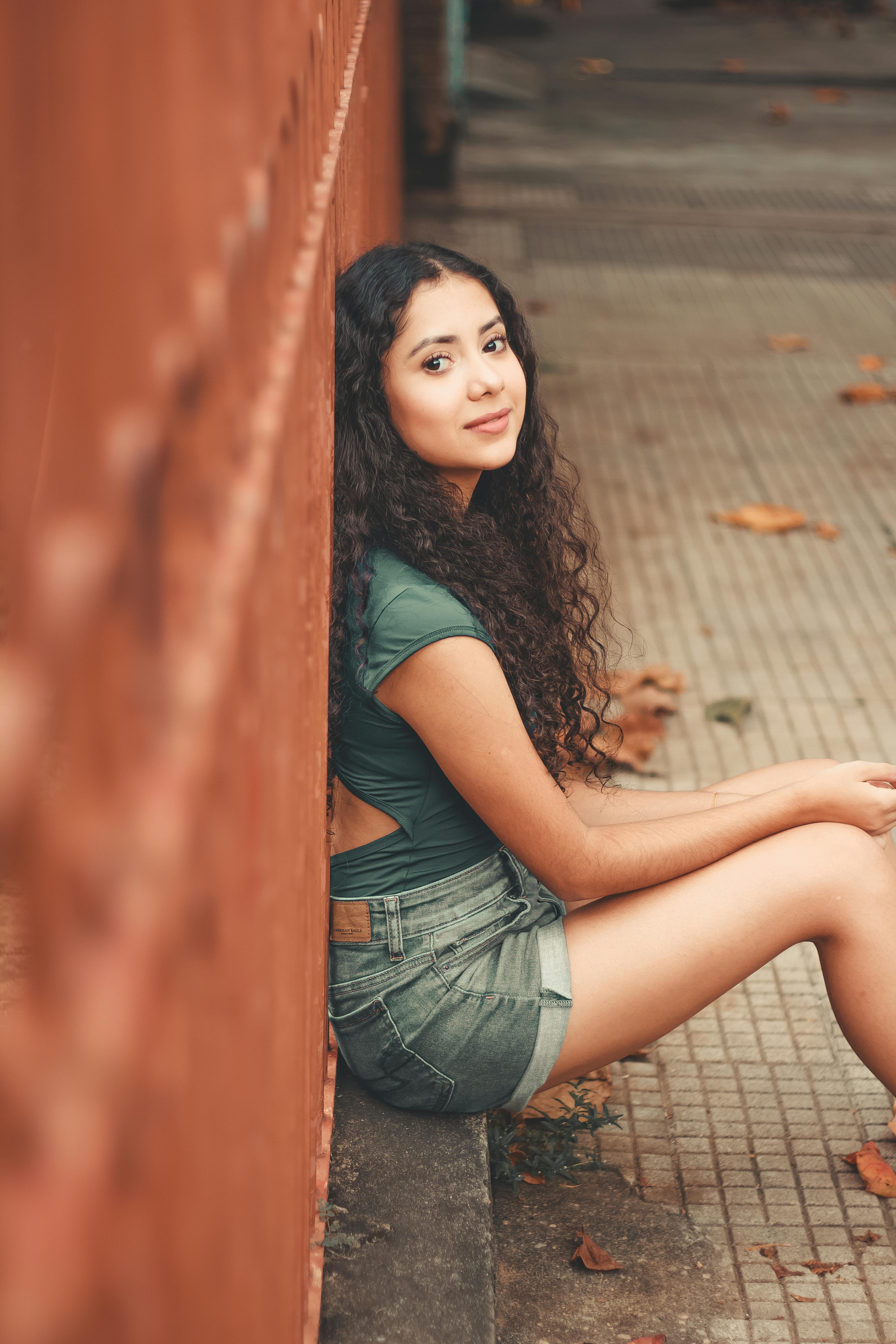 一位年轻卷发女子随意地坐在户外墙边，拍下这张肖像