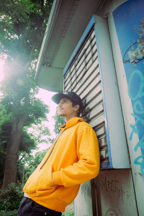 Free Stylish young man in an orange hoodie and black cap leaning against a wall outdoors, São Paulo. Stock Photo