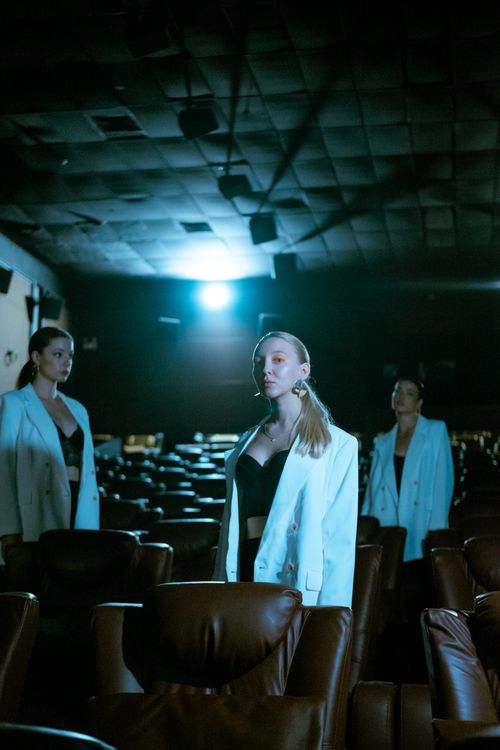 Free Three women in white jackets pose elegantly in a dimly lit theater, capturing fashion and style. Stock Photo