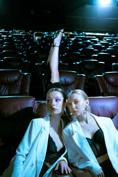 Free Chic fashion portrait of two women in white jackets posing in a cinema with dramatic lighting. Stock Photo