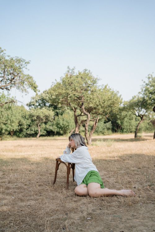 Free A woman leans on a chair in a sunlit meadow, surrounded by trees and tranquility. Stock Photo