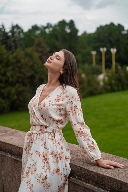 Free Elegant woman in a floral dress enjoying the outdoors with eyes closed, standing by a stone wall. Stock Photo