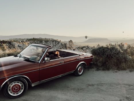 A vintage convertible car overlooking Cappadocia's unique rock formations and hot air balloons at sunset.
