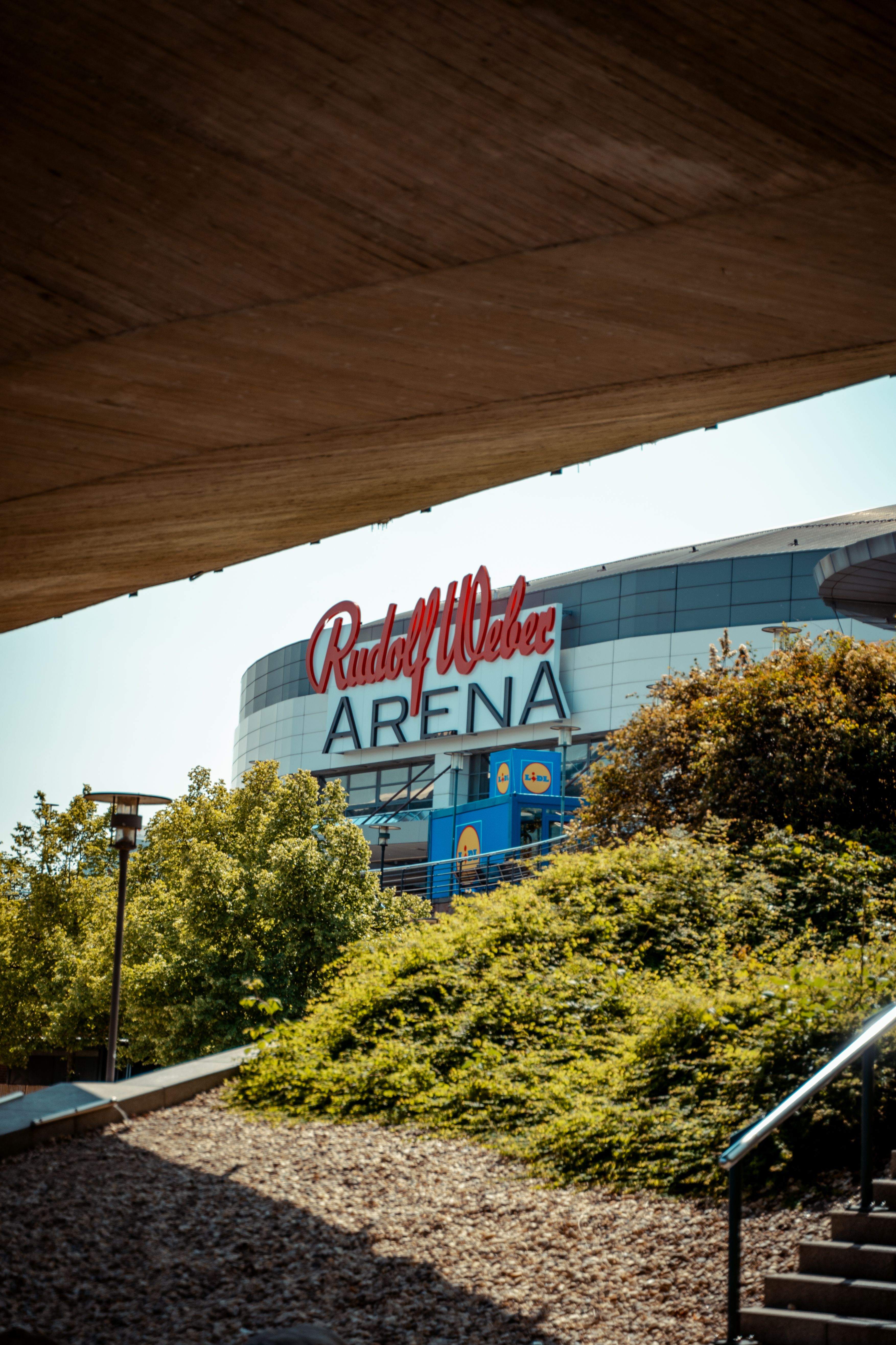 Vertical shot of Rudolf Weber Arena's modern facade in Oberhausen, Germany, surrounded by greenery.