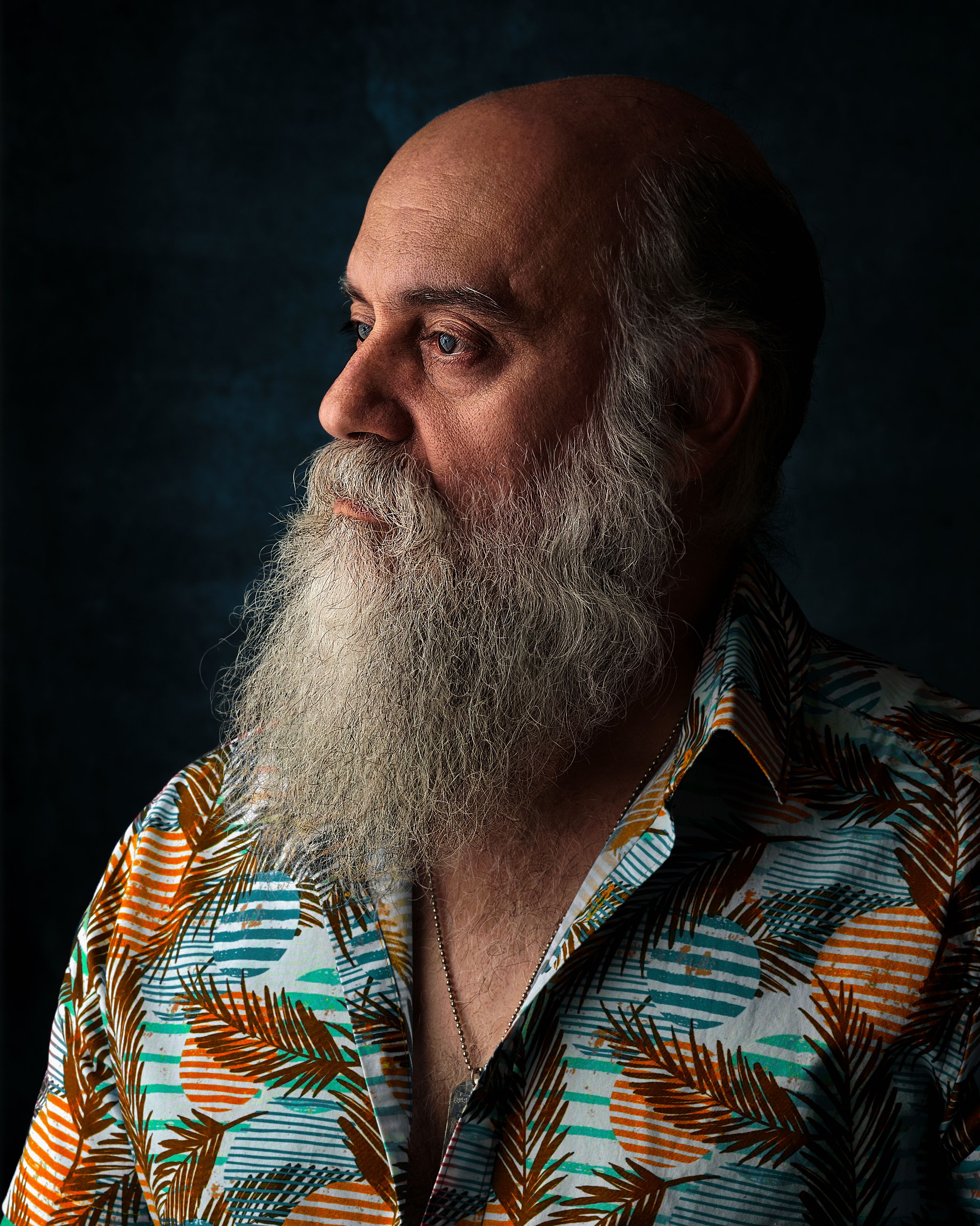 Portrait of an elderly man with a long white beard wearing a colorful shirt against a dark background.
