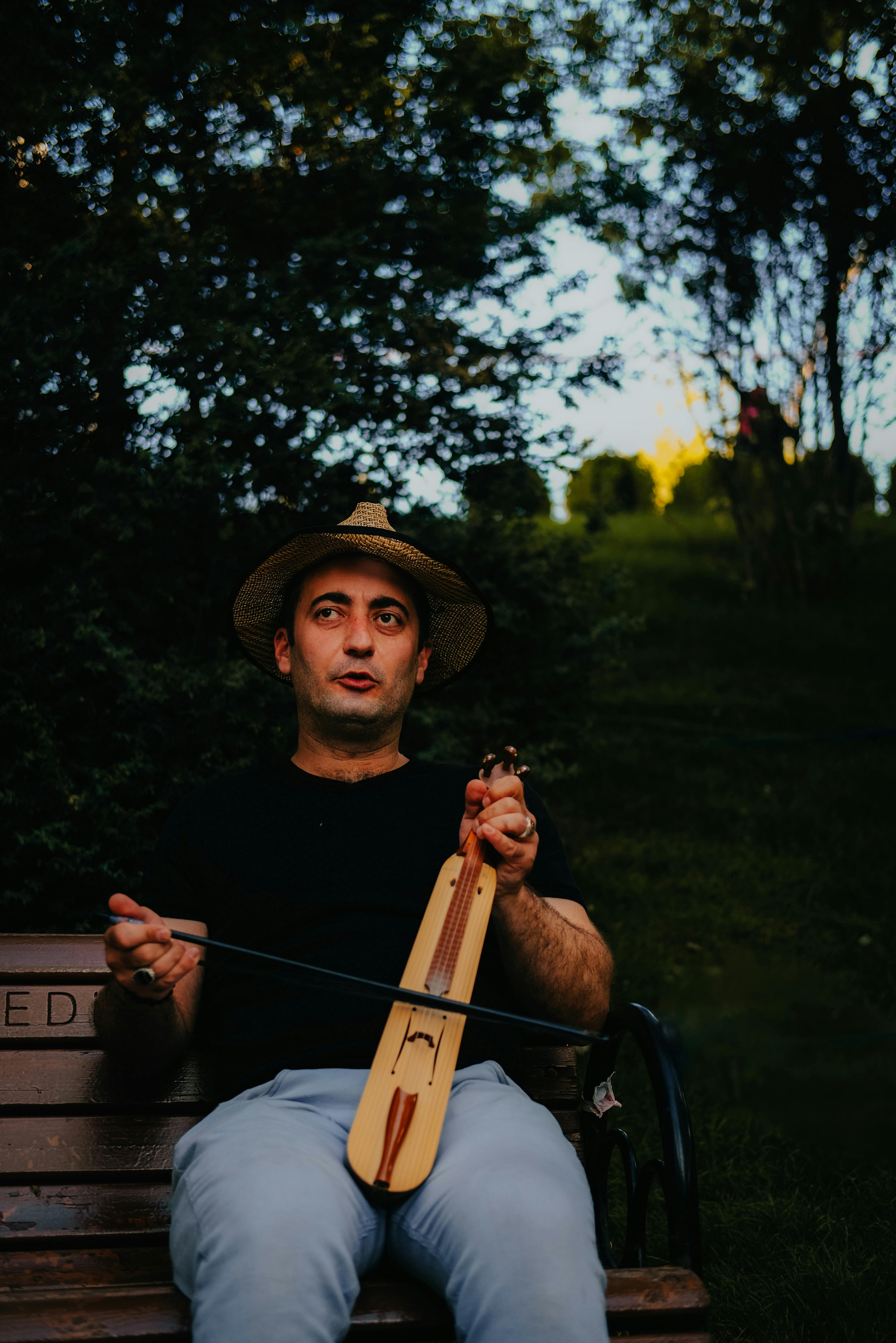 A man sitting on a bench in Ankara, playing a traditional Turkish string instrument under a hat.
