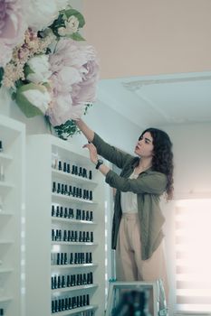 A young woman arranges large floral decorations in a cosmetics store setting with shelving and beauty products.