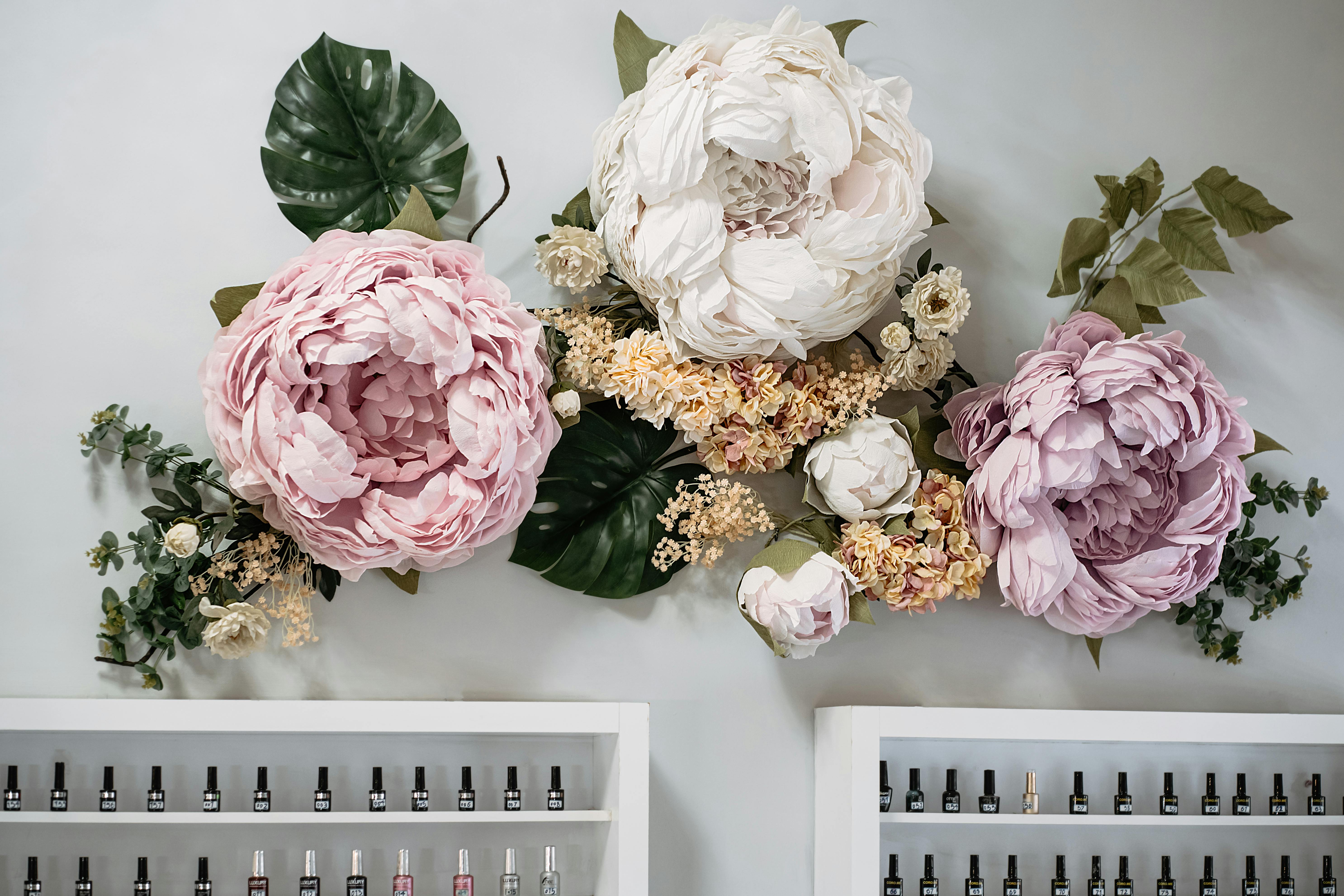 A beautiful arrangement of large artificial flowers adorning a wall above neat shelves with nail polish bottles.