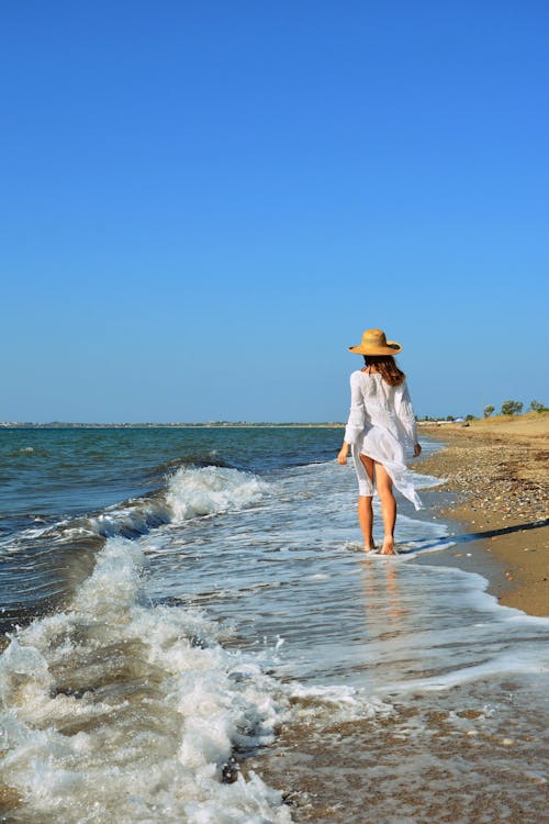 Woman in Hat Walking on Sea Shore under Clear Sky