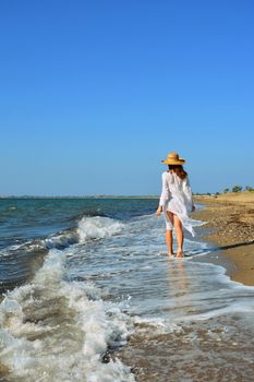 A woman in a hat and beachwear walks by the sea on a sunny day, enjoying a peaceful vacation.