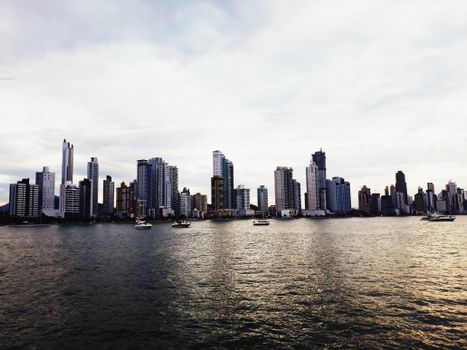 Amazing scenery of river with boats floating near tall modern glass skyscrapers in overcast weather