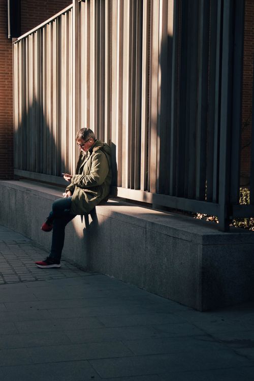 Free A man leans against a concrete wall in Madrid, Spain, using his smartphone on a sunny day. Stock Photo
