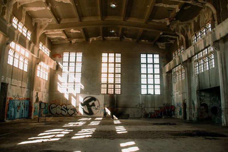 Sunlit interior of an abandoned industrial hall with graffiti and dramatic shadows.