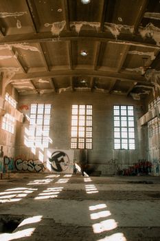 Sunlight streaming through windows of an abandoned warehouse with graffiti-covered walls.