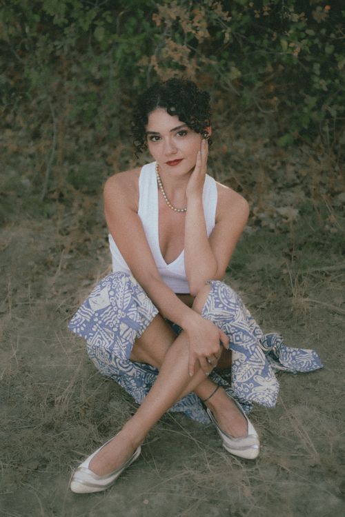 Free A stylish woman in a white tank and blue floral skirt sits gracefully outdoors. Stock Photo