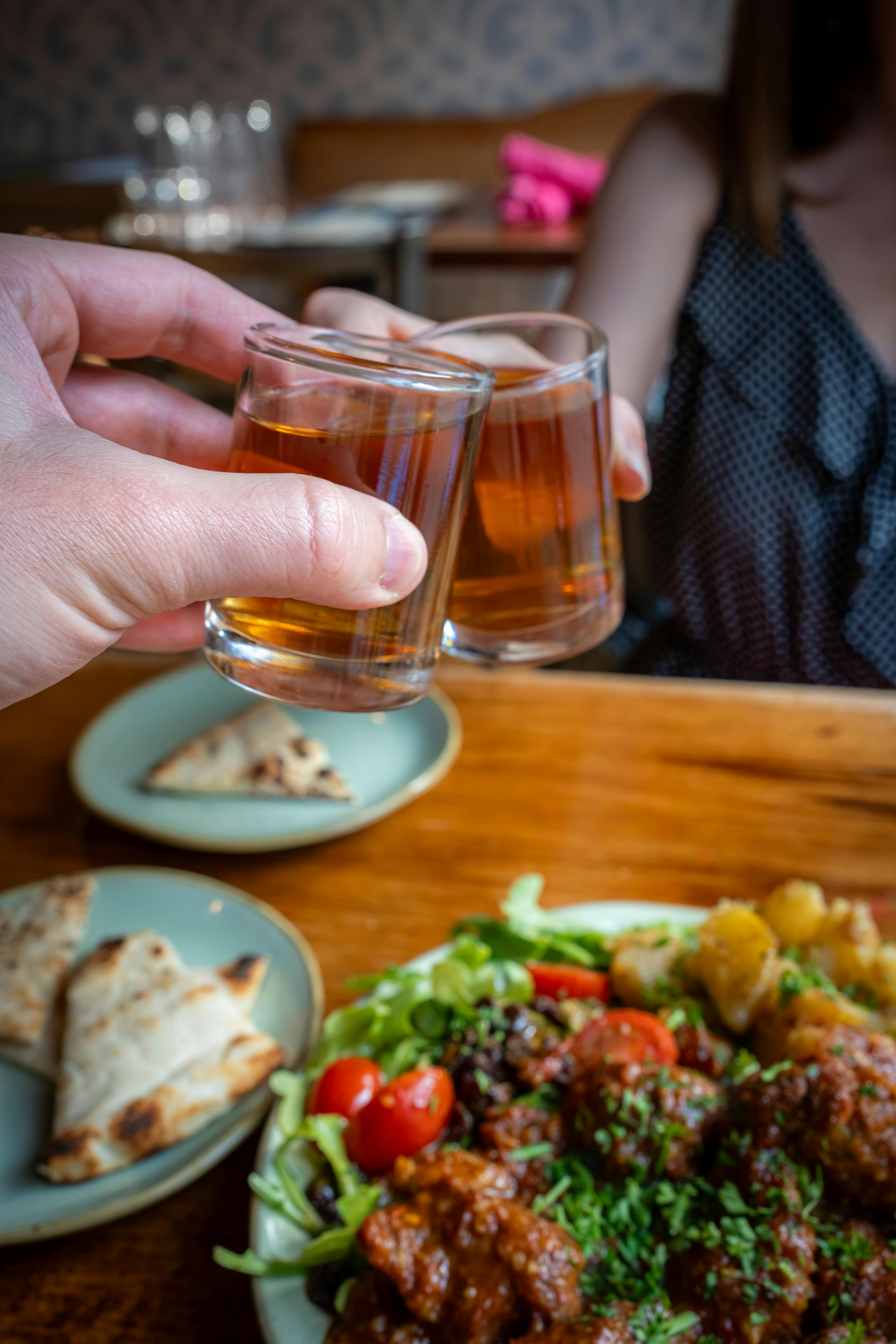 Two people toast with whiskey glasses over a hearty meal, showcasing dining and celebration.