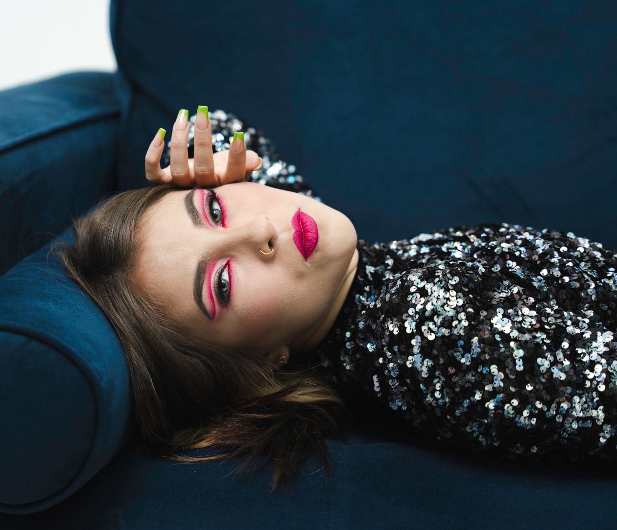 Woman with bold pink eye makeup and pink lipstick wearing a dark sequin top on a blue sofa