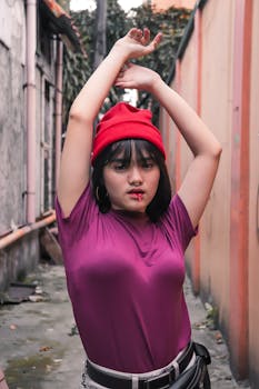 一位时尚的年轻女子头戴红色无檐帽，身着紫色上衣，在色彩缤纷的城市小巷里自信地摆出姿势